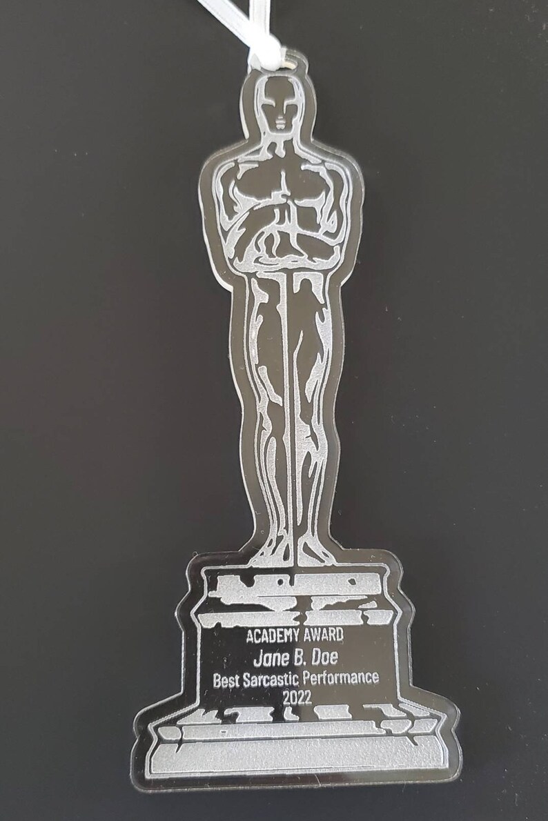 Puede incluir: Una estatuilla de Oscar de pl&aacute;stico plateado con una cinta blanca. La base de la estatuilla dice "ACADEMY AWARD Jane B. Doe Best Sarcastic Performance 2022".