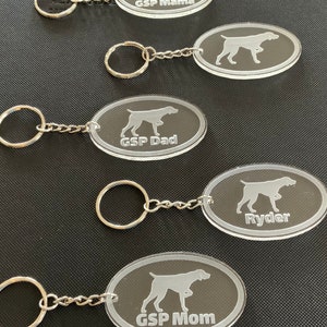 May include: Five clear oval-shaped keychains with silver key rings and chains. Each keychain features a silhouette of a dog and text. The keychains read "GSP Mama", "Ryder", "GSP Dad", and "GSP Mom".