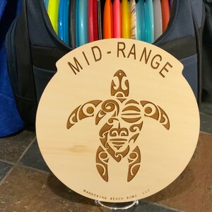 Op de afbeelding: Een houten schijf met een lasergesneden ontwerp van een schildpad met ingewikkelde patronen. De schijf is gelabeld met "MID-RANGE" en "WANDERING BEACH BUMS LLC".
