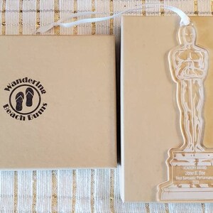 Puede incluir: Una caja marr&oacute;n con el texto "Wandering Beach Bunnies" y un par de chanclas dentro de un c&iacute;rculo. La caja est&aacute; al lado de un premio de acr&iacute;lico transparente con forma de persona sosteniendo un trofeo. El premio tiene el texto "Academy Award John E. Doe Mejor interpretaci&oacute;n dram&aacute;tica 2023" en la base.