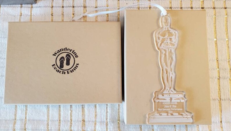 Puede incluir: Una caja marr&oacute;n con el texto "Wandering Beach Bunnies" y un par de chanclas dentro de un c&iacute;rculo. La caja est&aacute; al lado de un premio de acr&iacute;lico transparente con forma de persona sosteniendo un trofeo. El premio tiene el texto "Academy Award John E. Doe Mejor interpretaci&oacute;n dram&aacute;tica 2023" en la base.