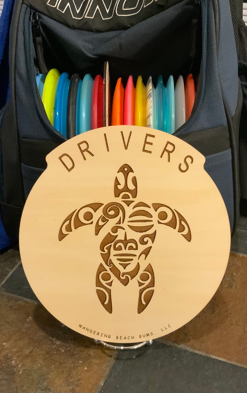 Op de afbeelding: Een houten schijf met een gesneden schildpadontwerp en de woorden "DRIVERS" en "WANDERING BEACH BUMS, LLC".