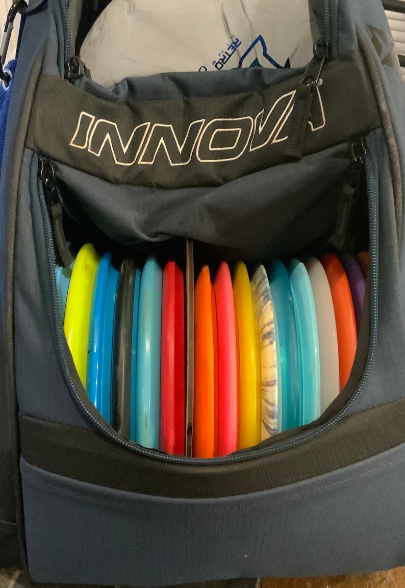 Op de afbeelding: Een blauwe discgolf-tas met het woord "INNO" erop gedrukt. De tas is open en gevuld met een verscheidenheid aan kleurrijke discgolf-schijven.