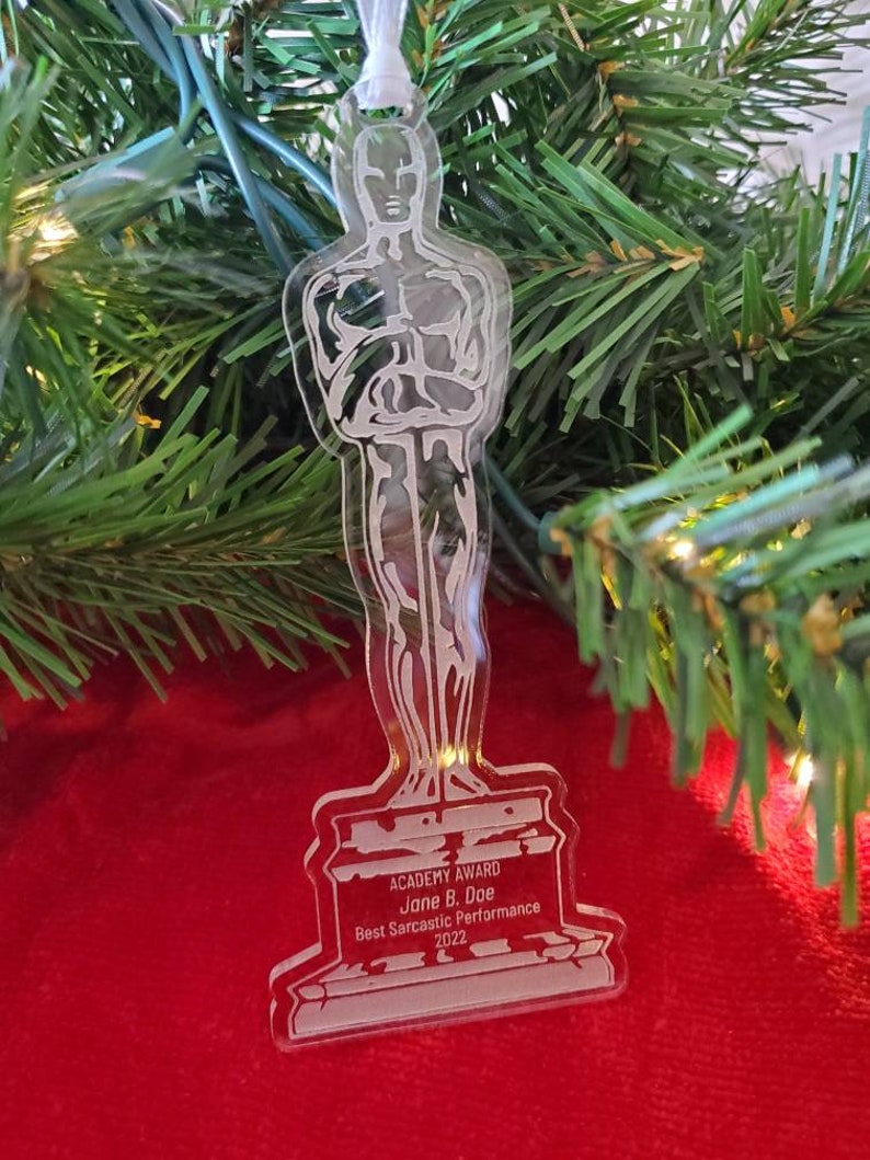 Puede incluir: Un adorno de pl&aacute;stico transparente con forma de la estatuilla del Premio de la Academia. La base del adorno tiene el texto "ACADEMY AWARD", "Jane B. Doe", "Best Sarcastic Performance" y "2022".