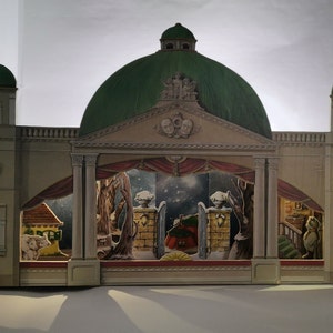 May include: A detailed paper theater stage with a green dome roof and beige facade. The stage features a winter scene with trees, a house, and figures. The design includes columns, arches, and a red curtain.