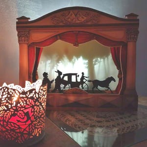 May include: A wooden shadow box with a red velvet curtain and a silhouette scene of a horse-drawn carriage in a forest. The shadow box is lit from behind, creating a dramatic effect.