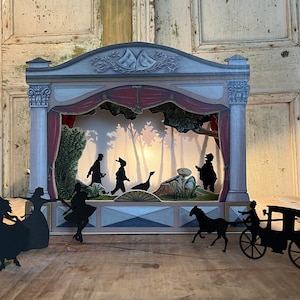 May include: A shadow puppet theater with a red curtain and a light shining through the back. The theater is made of cardboard and has a wooden base. There are five shadow puppets inside the theater, including a goose, a man with a hat, and a man with a beard. There are also four shadow puppets outside the theater, including two dancers and a horse-drawn carriage.