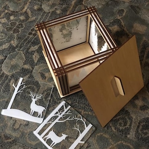 Op de afbeelding: Een houten doos met een scharnierend deksel, versierd met een winterscène. De doos is open en onthult een heldere glazen inzet met een winterscène. Twee aparte glazen stukken met een herten silhouet zijn ook inbegrepen.