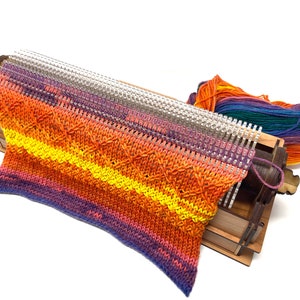May include: A wooden knitting loom with a colorful knitted scarf in shades of purple, orange, and yellow. The loom is made of wood and has a metal needle. The scarf is partially knitted and is hanging off the loom.