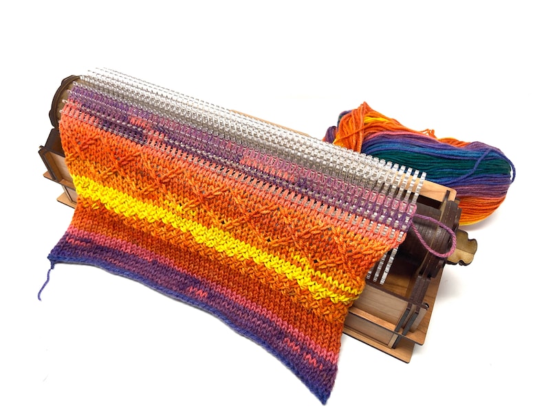 May include: A wooden knitting loom with a colorful knitted scarf in shades of purple, orange, and yellow. The loom is made of wood and has a metal needle. The scarf is partially knitted and is hanging off the loom.