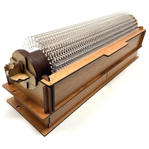 May include: A wooden music box with a clear plastic comb and a brown wooden crank. The music box is made of light wood with dark brown accents.