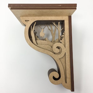 Puede incluir: Escuadra de estante decorativa de madera con un recorte de escena de bosque. El soporte presenta un ciervo de pie entre árboles y colinas. El estante está hecho de madera marrón claro con un borde marrón oscuro y un diseño decorativo de volutas.