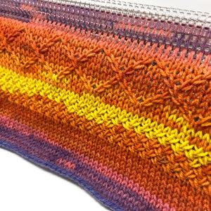 May include: A close-up of a knitted fabric with a multi-colored ombre pattern in shades of purple, orange, and yellow. The fabric is being woven on a loom.