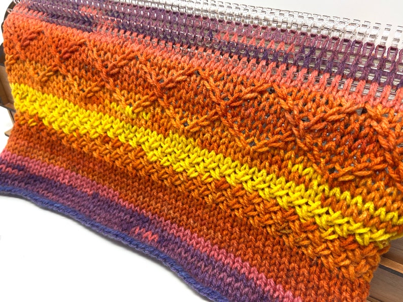 May include: A close-up of a knitted fabric with a multi-colored ombre pattern in shades of purple, orange, and yellow. The fabric is being woven on a loom.