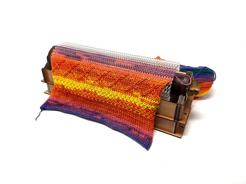 May include: A colorful knitted fabric is being woven on a wooden loom. The fabric is a mix of orange, yellow, and purple yarn. A ball of rainbow yarn is sitting next to the loom.
