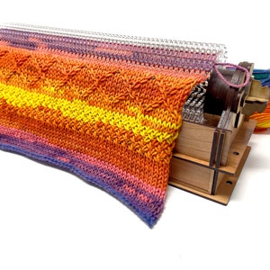 May include: A wooden knitting loom with a colorful knitted fabric on it. The loom has a clear plastic needle and a wooden crank. The fabric is a mix of orange, yellow, and purple.