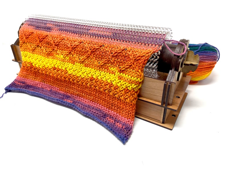 May include: A wooden knitting loom with a colorful knitted fabric on it. The loom has a clear plastic needle and a wooden crank. The fabric is a mix of orange, yellow, and purple.