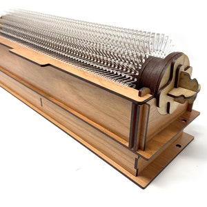 May include: A wooden music box with a clear plastic comb and a brown wooden crank. The music box is made of light wood with dark brown accents.
