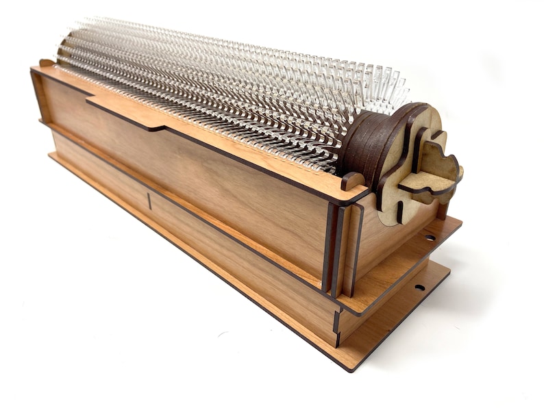 May include: A wooden music box with a clear plastic comb and a brown wooden crank. The music box is made of light wood with dark brown accents.
