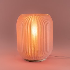 May include: A pink, ribbed-textured table lamp with a warm, glowing light emanating from within. The lamp has a cylindrical shape and sits on a light-colored base. A white power cord extends from the base.