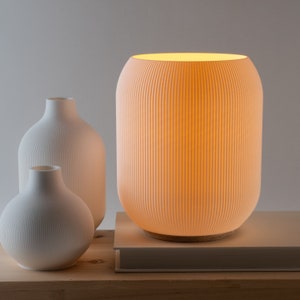 May include: A white ceramic table lamp with a ribbed design and a warm white light. The lamp is on a white book on a wooden surface. There are two smaller white ceramic vases with ribbed designs on the surface.