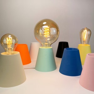 May include: A set of seven table lamps with different colored bases. The lamps have a variety of shapes and sizes, and they are all lit with warm white light bulbs. The lamps are arranged in a row on a white surface.
