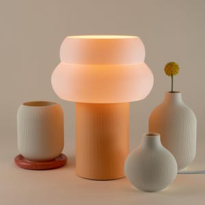May include: A white ceramic table lamp with a double-layered shade and a ribbed orange base. The lamp is lit and casting a warm glow. There are three white ceramic vases with ribbed textures on the table, one with a yellow flower.