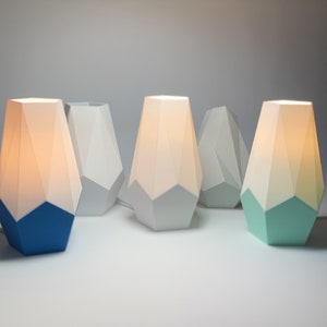 May include: Five geometric table lamps with white and colored bases. The lamps are lit and have a warm glow. The bases are white, blue, and green.