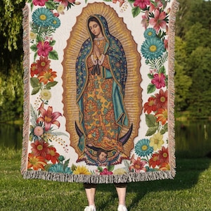 May include: A woven throw blanket featuring a central image of Our Lady of Guadalupe surrounded by a vibrant floral border. The design includes a golden sunburst, a blue mantle with stars, and a yellow dress, all framed by colorful flowers.