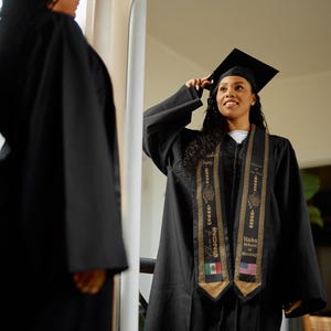 May include: A person in a black graduation gown and cap, adjusting the cap. The gown has a gold and black sash with the text "Hahn School of Nursing" and the year "2023". The sash also features the flags of Mexico and the United States.