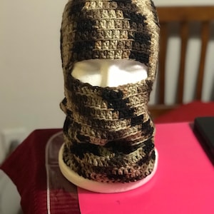 May include: A brown, black, and tan camouflage crochet balaclava with a face opening. The balaclava is on a white mannequin head.