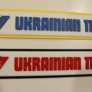 May include: Two rectangular signs with black and yellow borders. The top sign has a blue Ukrainian coat of arms and the text "UKRAINIAN TIRE" in blue. The bottom sign has a red Ukrainian coat of arms and the text "UKRAINIAN TIRE" in red.