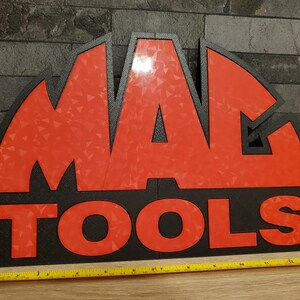 May include: A black and red sign with the text "MAC TOOLS" in a bold, white font. The sign is made of a textured material and has a slightly curved shape.