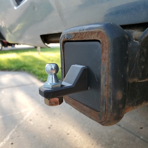 Pode incluir: Close-up de um engate de reboque de metal preto com uma bola prateada. O engate está montado em uma superfície de metal enferrujada.