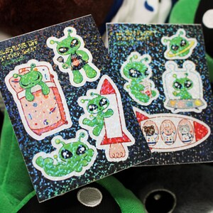 Op de afbeelding: Twee vellen holografisch stickerpapier met verschillende stickers met een alien-thema. De stickers tonen aliens in verschillende poses, waaronder slapen, vliegen in een raket en muziek maken. De tekst op het stickervel luidt "Blesstings Off Sticker Sheet".