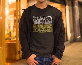 May include: A man wearing a black t-shirt with a yellow and black graphic that says "Night Kids" and "Myogi Racing Team" in Japanese. The man is standing in front of a shop with a sign that says "A016."