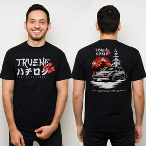 May include: Black t-shirt featuring Japanese text and a red floral design on the front. The back of the shirt displays a car graphic with Japanese text, a red sun, and mountains. AE86 (Hachi-Roku) is printed on the front.