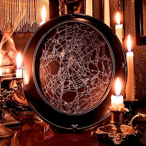 May include: A dark, moody image featuring a round, black framed decorative tray with a white spiderweb design. Lit candles of varying heights and styles surround the tray, creating a warm, flickering glow. The scene evokes a Halloween or gothic theme.
