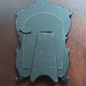 May include: Black picture frame with an ornate design and a stand on the back. The frame is made of cardboard and has a textured surface.