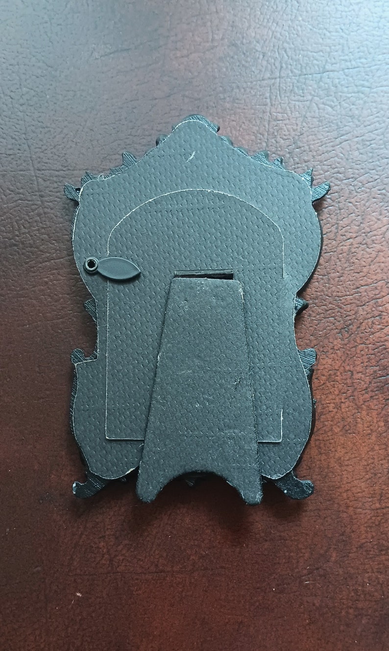 May include: Black picture frame with an ornate design and a stand on the back. The frame is made of cardboard and has a textured surface.