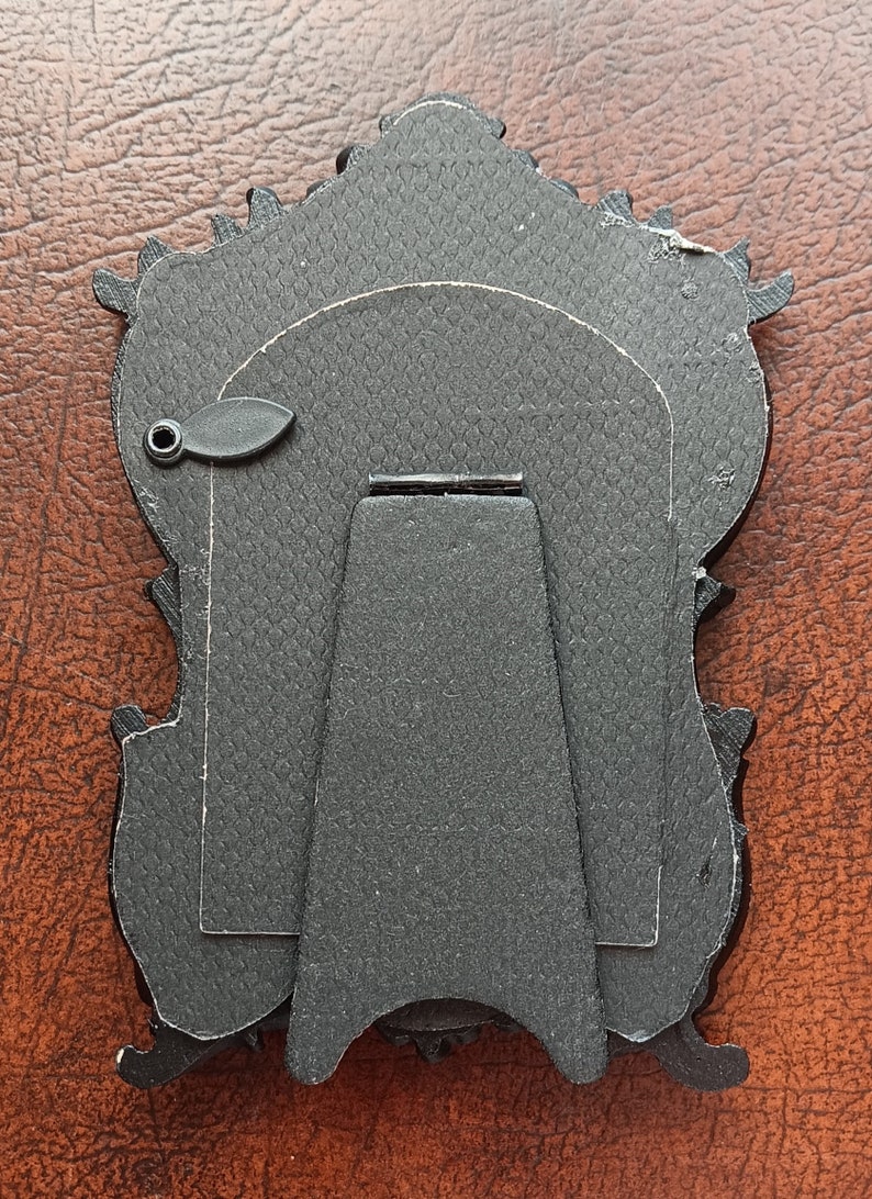 May include: Black ornate picture frame with a stand on the back. The frame has a textured surface and a small metal loop on the side.