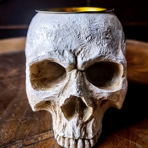 Pode incluir: Queimador de incenso de caveira de cer&acirc;mica branca com topo de metal dourado. A caveira tem um design detalhado e est&aacute; sobre uma superf&iacute;cie de madeira.