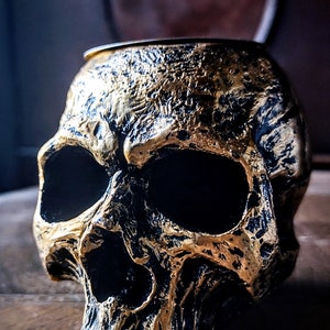 Pode incluir: Um porta-velas em forma de caveira dourado e preto. A caveira &eacute; detalhada com caracter&iacute;sticas texturizadas e uma &oacute;rbita ocular preta.