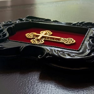 May include: A gold cross with intricate details is displayed in a black ornate frame with a red velvet backing.