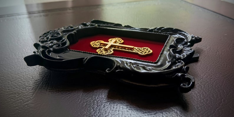 May include: A gold cross with intricate details is displayed in a black ornate frame with a red velvet backing.