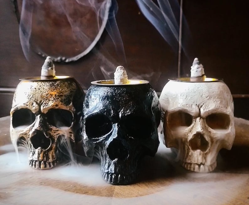 Pode incluir: Tr&ecirc;s queimadores de incenso em forma de caveira, um dourado, um preto e um branco, com varetas de incenso a arder no seu interior.