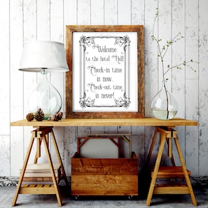 May include: A framed print with the text "Welcome to the hotel Hell! Check-in time is now. Check-out time is never!" in a decorative font. The frame is brown, and the print is displayed on a wooden table with a lamp and vase.