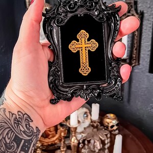 May include: A black ornate frame with a gold cross inside. The frame is held in a hand with a tattoo.