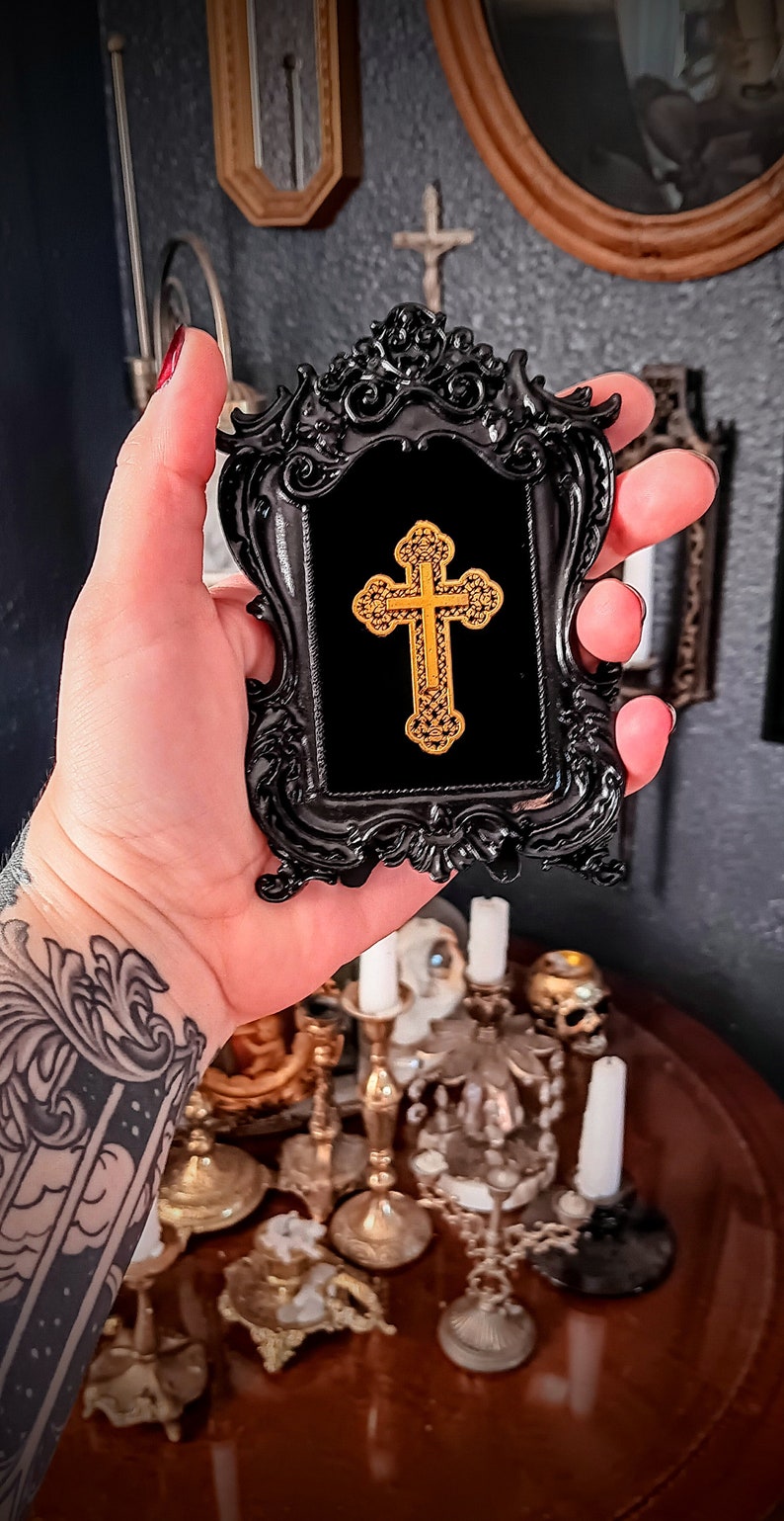 May include: A black ornate frame with a gold cross inside. The frame is held in a hand with a tattoo.
