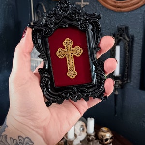 May include: A black ornate frame with a red velvet mount and a gold cross inside. The frame is held in a hand with a tattoo.